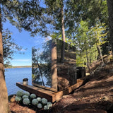 Custom Outdoor Wellhouse IceCube sauna wrapped in mirror that is reflecting majestic lakeside scene.  Beautiful Muskoka Lakeview with towering trees and blue sky are all visible while looking into the mirrored exterior of the sauna.